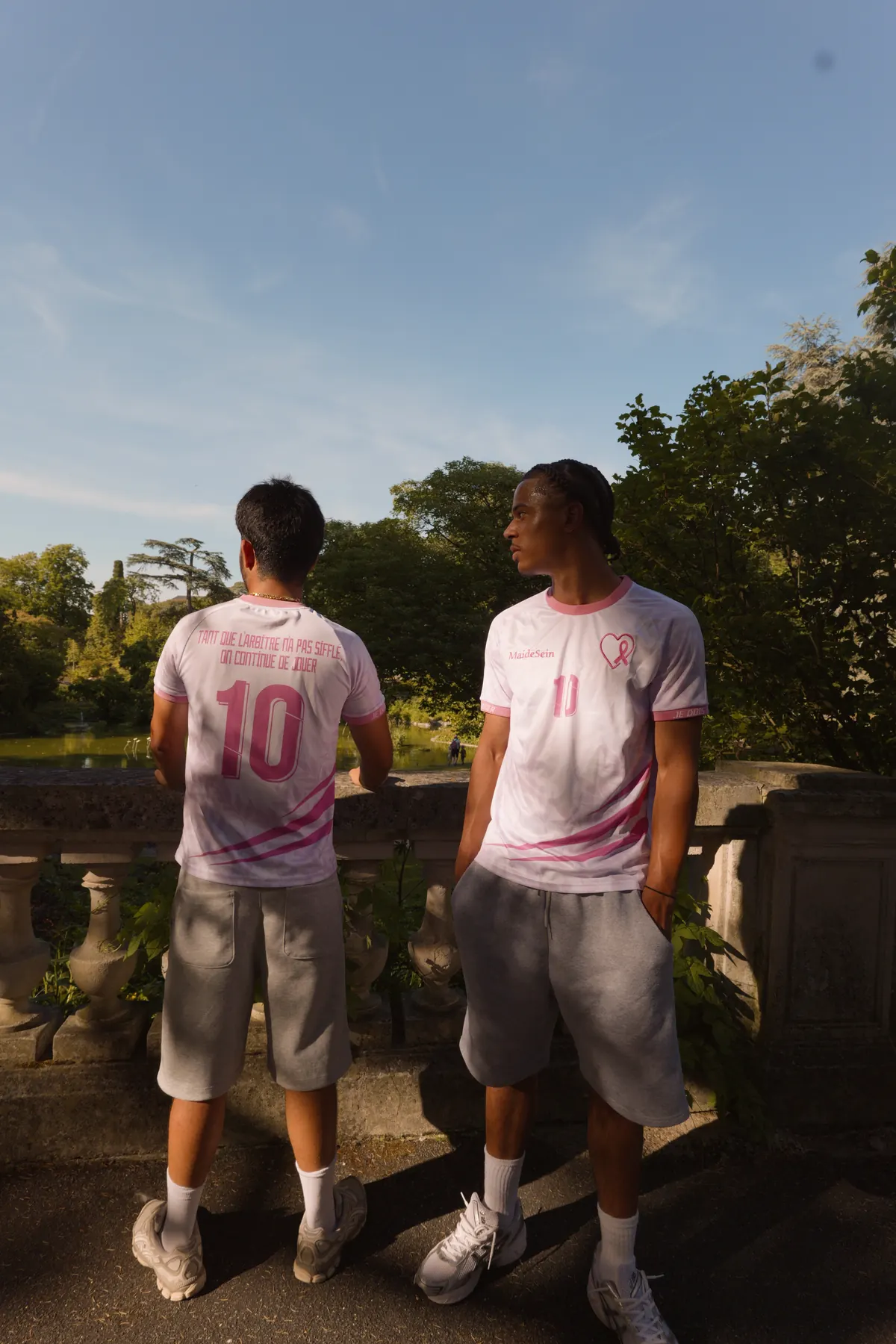 Dans un parc, deux jeunes hommes portent le maillot MAIDESEIN blanc et rose ; l’un, de dos, montre le numéro 10 et la signature de l’association