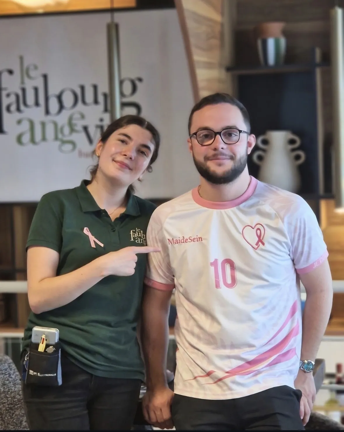 Au Faubourg Angevin, une serveuse portant le ruban rose désigne en souriant le maillot solidaire MAIDESEIN porté par un homme, devant le logo de la brasserie