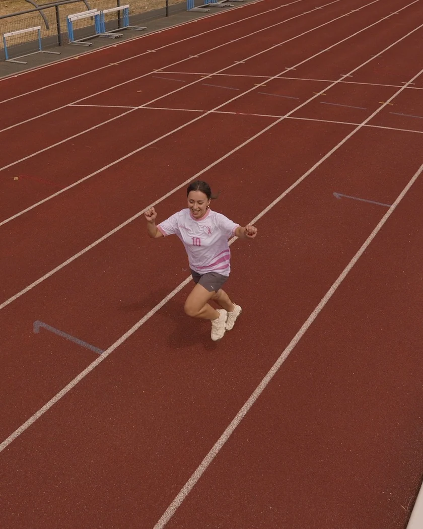 Sur une piste d’athlétisme, une jeune femme en maillot MAIDESEIN court en souriant, les poings levés